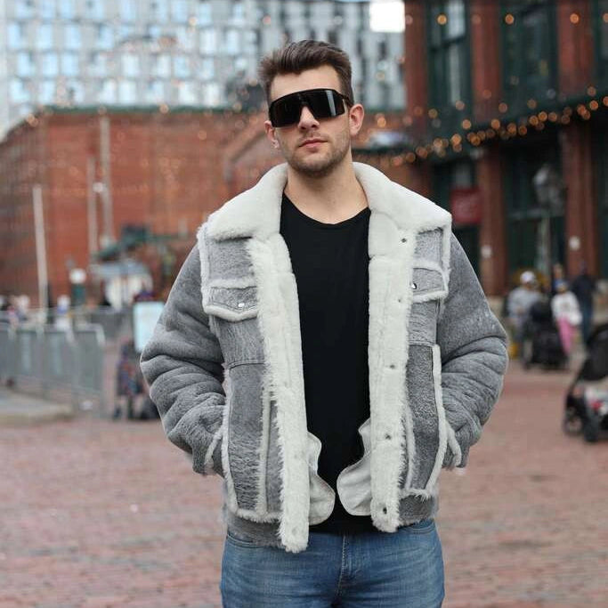 Man wearing sunglasses and a distressed grey shearling jacket with white fur trim in an urban setting.