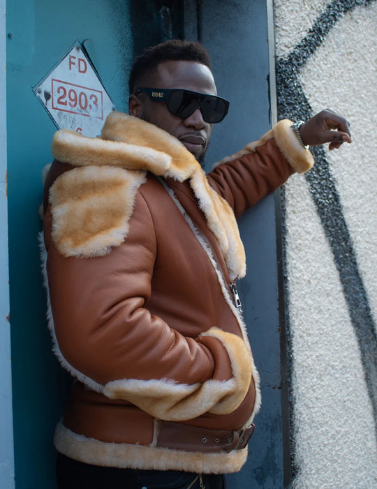 Alek's Biker Shearling Jacket with Fur Details, shown worn by a man standing against a blue door, featuring a zipper front and fur-lined collar and cuffs. The jacket is styled with a brown leather body and beige fur accents.