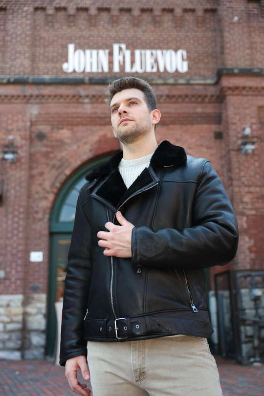Man wearing Black on Black Belted Shearling Leather Biker Jacket in front of a brick building with 'John Fluevog' sign.