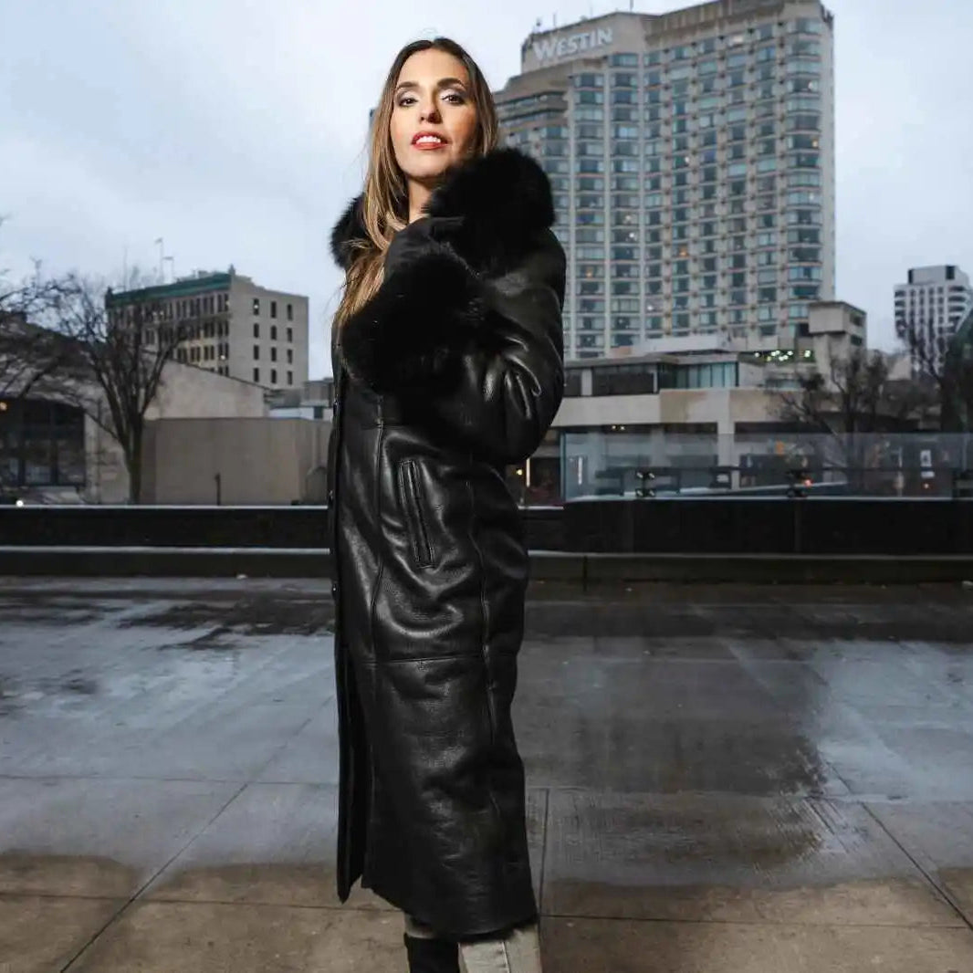 Woman in a black shearling sheepskin coat standing with cityscape in the background.